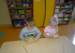 Dwoje dzieci siedzi przy stole, w rękach trzymają gumki recepturki, które zakładają na kartonowe małe pudełko.